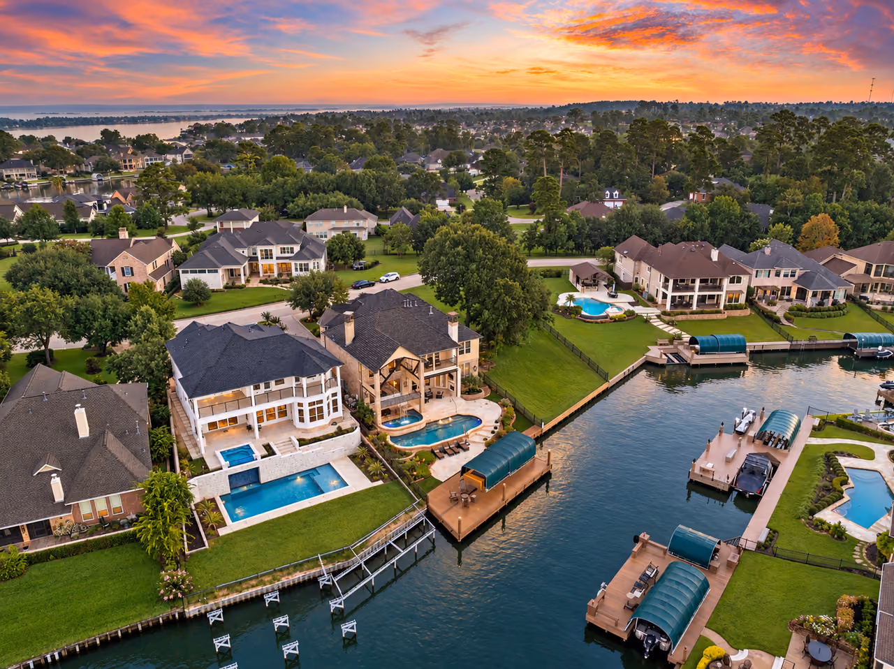 Aerial view at sunset over large two-story houses on a channel: lit pools and stone terraces run down to the bank, and wooden docks hold boat slips under green arched covers. Beyond them, more houses stand among tall pines and oaks, and open water reaches the horizon under an orange and pink sky.